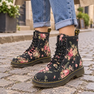 May include: A pair of black lace-up ankle boots featuring a floral pattern with pink and cream roses and green leaves. The boots have black laces, a black sole with yellow stitching, and a yellow heel pull tab with black text on the back.
