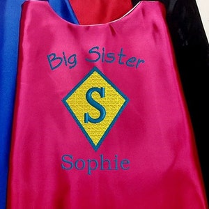 May include: Pink superhero cape with the words "Big Sister" and "Sophie" embroidered on it. A yellow diamond with a blue "S" is in the center.