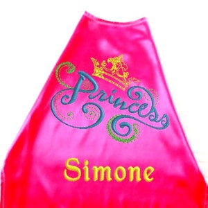 May include: Pink satin cape with a gold crown and the word "Princess" embroidered in blue and green. The name "Simone" is embroidered below in gold.