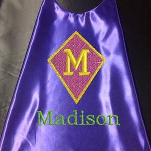 May include: Purple satin superhero cape with a pink diamond-shaped patch embroidered with the letter "M" in yellow thread. The name "Madison" is embroidered in green thread below the patch.