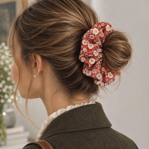 Peut inclure: Un chouchou en tissu avec un fond rouge vif orné d'un motif répétitif de petites fleurs blanches et jaunes ressemblant à des marguerites. L'accessoire est froncé en une forme circulaire et est porté autour d'un chignon de cheveux châtain clair.