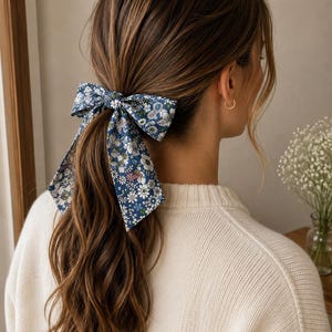 Peut inclure: Un nœud pour cheveux bleu orné d'un motif floral dense composé de fleurs blanches, bleu clair et roses avec des feuilles vertes. Le nœud est attaché autour d'une queue-de-cheval, avec deux longs pans de tissu pointus tombant sur les cheveux.