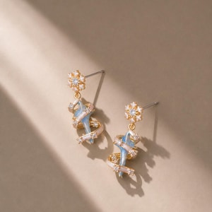May include: A pair of gold-tone drop earrings featuring a flower-shaped stud with a central clear stone. A light blue, sword-like element is wrapped in a spiraling, pale pink ribbon adorned with small, clear, sparkling stones along its length.