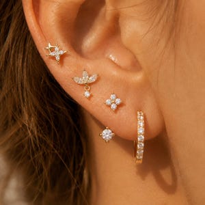 Könnte beinhalten: Ein Set aus fünf goldfarbenen Ohrringen an einem Ohr. Die Kollektion umfasst einen sternförmigen Stecker, einen floralen Stecker mit Marquise-Schliff, einen viersteinigen Cluster-Stecker, einen runden Solitär-Stecker und einen kleinen Creolen-Ohrring.