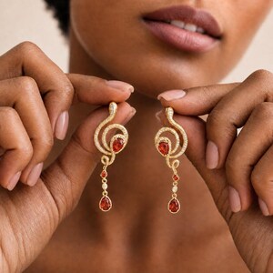May include: A pair of gold-toned dangle earrings featuring a swirling, textured metal design encrusted with small clear stones. Each earring is accented with two teardrop-shaped red gemstones and a single small clear stone set in the center.