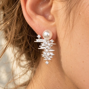May include: A silver-toned earring featuring a single round white pearl at the top, accented by a cluster of small, clear, sparkling stones arranged in a jagged, layered, and asymmetrical geometric pattern extending downward from the pearl.