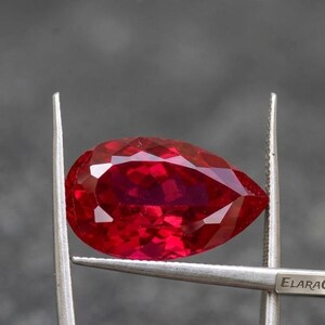 以下が含まれることがあります： 銀色の金属製ピンセットで保持された、洋ナシ型のファセットカットが施された赤い宝石。この石は、深く鮮やかな赤色をしており、光を反射する多数の三角形やひし形のファセットが、磨き上げられた半透明の表面全体に広がっています。