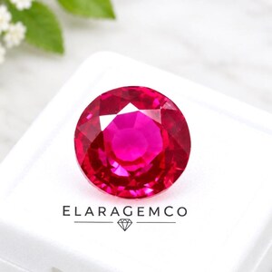 Pode incluir: Uma pedra preciosa redonda, facetada e de um vermelho vibrante com corte brilhante, exibindo tons carmesim profundos e reflexos de luz. A pedra está posicionada sobre uma superfície branca com o texto preto ELARAGEMCO e um logótipo de diamante.