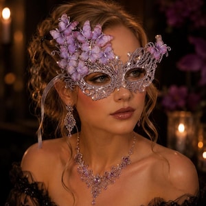 Op de afbeelding: Een zilveren filigraan masker voor een gemaskerd bal, versierd met paarse vlinders en heldere kristallen, gecombineerd met een bijpassende zilveren filigraan ketting en bungelende oorbellen met paarse stenen en heldere kristallen.