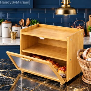 May include: A two-tier bamboo bread box with a light wood finish and a clear, ribbed glass drop-down door featuring a gold-toned handle. The interior shelves hold several baked goods, and the unit is constructed with visible metal screws.