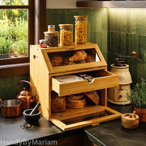 May include: A bamboo bread box with a clear front window, a middle storage drawer, and a bottom drop-down door. The unit holds various baked goods, including croissants and loaves of bread, and is topped with three glass canisters filled with dry pasta.