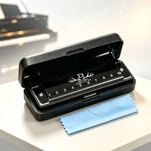 Könnte beinhalten: Eine schwarze Mundharmonika mit zehn nummerierten Löchern und einer individuellen Gravur mit Geweih und dem Text "Dylan 2026.04.25" liegt in einem offenen schwarzen rechteckigen Etui, begleitet von einem kleinen hellblauen Reinigungstuch.