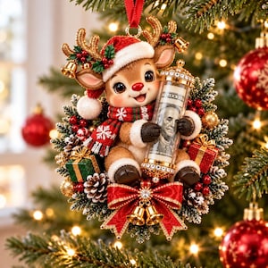 May include: A Christmas tree ornament featuring a cartoon reindeer wearing a red Santa hat and a red and green scarf. The reindeer holds a clear cylindrical tube containing a rolled-up one hundred dollar bill, surrounded by a wreath of pine, red berries, pinecones, and small gift boxes.