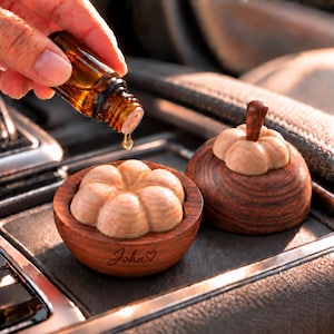 May include: A dark brown wooden essential oil diffuser with a pumpkin-shaped top and a matching base. The base features the engraved name John followed by a heart symbol. A small amber glass dropper bottle is dispensing a single oil drop.