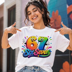 Op de afbeelding: Een wit T-shirt met korte mouwen en een kleurrijke afbeelding van de cijfers 6 en 7 met cartoongezichten, armen en handen. Onder de cijfers staat de tekst Six Seven in een vet, meerkleurig lettertype, omringd door kleine sterren en spatten.