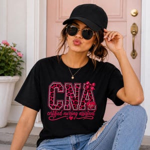 Könnte beinhalten: Ein schwarzes T-Shirt mit dem Text CNA in großen, pinkfarbenen Leopardenmuster-Buchstaben, die mit weißen Punkten umrandet sind. Eine kleine pinke Schleife und ein Herz mit EKG-Linie sind am Buchstaben A befestigt. Darunter steht certified nursing assistant.