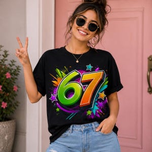 Op de afbeelding: Een zwart t-shirt met korte mouwen voorzien van een grote, levendige afbeelding van het getal 67 in felgroen en oranje. Het ontwerp is omringd door kleurrijke abstracte spatten, verfspatten en diverse stervormen in tinten paars, blauw en groen.