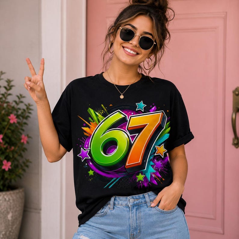 Op de afbeelding: Een zwart t-shirt met korte mouwen voorzien van een grote, levendige afbeelding van het getal 67 in felgroen en oranje. Het ontwerp is omringd door kleurrijke abstracte spatten, verfspatten en diverse stervormen in tinten paars, blauw en groen.