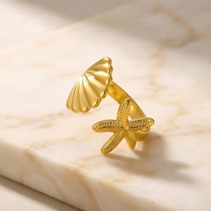 Può includere: Un anello aperto color oro caratterizzato da un motivo a conchiglia lucida su un'estremità e un design a stella marina testurizzata a cinque punte sull'altra. La fascia è liscia e curva per collegare i due elementi marini in un anello.