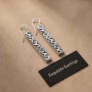 May include: A pair of silver drop earrings featuring a long rectangular bar design with intricate embossed scrollwork and floral patterns. The earrings include curved ear wires and are displayed next to a black card with the text Exquisite Earrings.