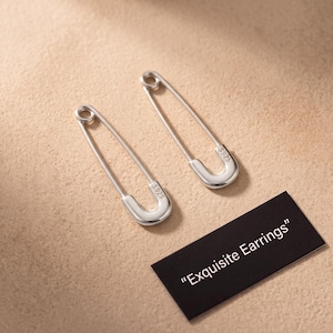 May include: A pair of silver-toned safety pin earrings with a polished finish. Each earring features the engraving S925 on the clasp. A small black rectangular card is present with the white printed text "Exquisite Earrings".