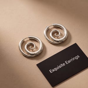 Pode incluir: Um par de brincos de argola prateados com um design de espiral central. Ao lado dos brincos, há um cartão retangular preto com o texto branco Exquisite Earrings. Os brincos possuem um acabamento metálico polido e liso.