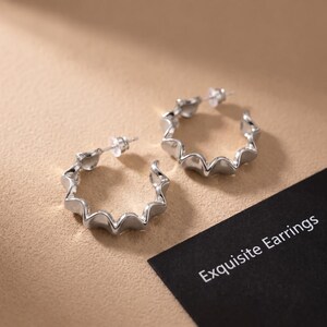 May include: A pair of silver-toned hoop earrings with a wavy, scalloped edge design and clear plastic earring backs. A black card with the white text Exquisite Earrings is positioned partially underneath the jewellery pieces.