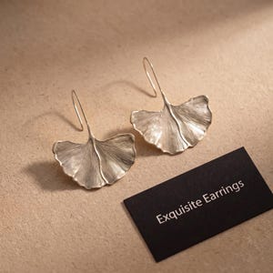 Pode incluir: Um par de brincos prateados em forma de folhas de ginkgo com texturas de linhas delicadas e radiantes e ganchos curvos. Um pequeno cartão retangular preto está posicionado próximo com o texto impresso em branco Exquisite Earrings.