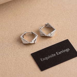 May include: A pair of silver-toned hoop earrings with a geometric, angular pentagonal shape. The earrings feature a hinged post mechanism. Next to the earrings is a small black rectangular card with the white printed text Exquisite Earrings.