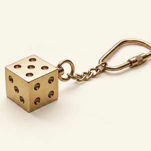 Puede incluir: Un llavero con forma de dado cúbico en tono dorado metálico con puntos circulares hundidos en sus caras. El dado está unido a una cadena metálica corta conectada a un mosquetón ovalado a juego con un mecanismo de cierre roscado.