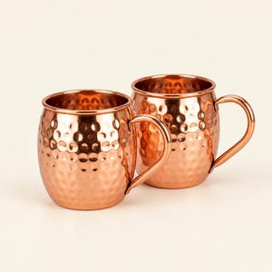 Peut inclure: Deux tasses rondes en cuivre présentant une texture martelée distincte sur toute la surface extérieure. Chaque tasse est équipée d'une anse lisse et incurvée fixée sur le côté, avec une finition métallique polie et un ton cuivre chaud.