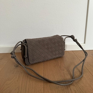 Könnte beinhalten: Eine taupefarbene, rechteckige Umhängetasche mit einem geflochtenen Ledermuster auf der Außenseite. Die Tasche verfügt über einen passenden, dünnen Kordelriemen mit Knoten an beiden Seiten, der durch integrierte Schlaufen befestigt ist.