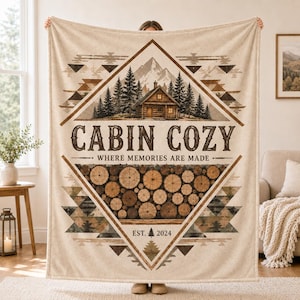 May include: A beige throw blanket featuring a diamond-shaped graphic with a cabin in a forest, a stack of logs, and the text "CABIN COZY WHERE MEMORIES ARE MADE" and "EST. 2024". The design includes geometric patterns in brown and tan tones.