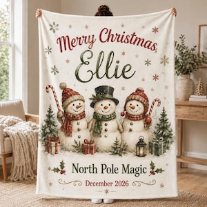 May include: A cream-coloured fleece blanket featuring the text Merry Christmas, Ellie, and North Pole Magic, December 2026. The design includes three snowmen, two wearing red hats and scarves, one wearing a black top hat, surrounded by pine trees, gifts, and snowflakes.