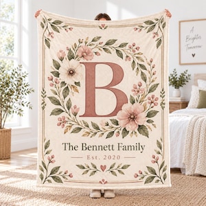May include: A cream-coloured throw blanket featuring a large, muted pink letter B centred within a circular wreath of soft pink flowers and green leaves. Below the wreath, the text reads The Bennett Family and Est. 2020 with a small heart icon.