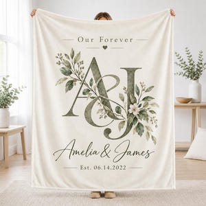 May include: A cream-coloured throw blanket featuring the text Our Forever, the large monogram letters A and J, the names Amelia & James, and the date Est. 14.06.2022. The design includes watercolour-style green leaves, branches, and a white flower.