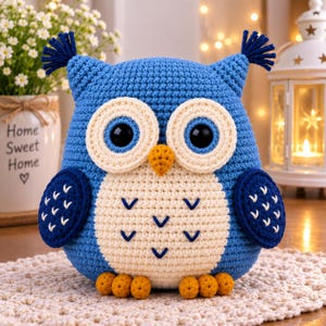 Puede incluir: Un peluche de búho tejido a ganchillo en color azul con grandes ojos negros, discos faciales color crema y un pico amarillo. El búho presenta mechones en las orejas, alas azul oscuro con bordados en V blancos y seis pies amarillos.