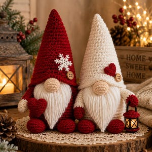 May include: Two crochet gnomes with tall pointed hats. The left gnome has a red hat with a white snowflake and a wooden button, holding a red heart. The right gnome has a cream hat with a red heart and a wooden button, holding a small red lantern.