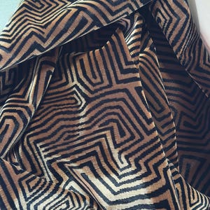 May include: A length of fabric featuring a repeating geometric maze pattern in shades of black and tan. The material has a soft, textured pile surface with deep folds that create shadows and highlight the intricate, angular lines of the design.