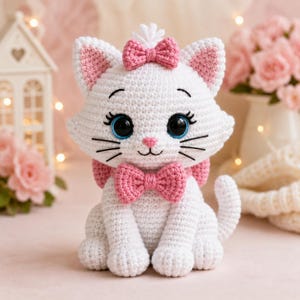 May include: A white crocheted cat plush toy featuring large blue eyes, a small pink nose, and black embroidered whiskers. The cat is adorned with a pink crocheted bow on its head and a matching pink bow around its neck, with pink inner ears.