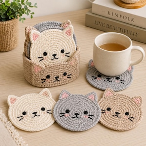 Può includere: Un set di sottobicchieri rotondi all'uncinetto a forma di gatto nei colori crema, grigio e beige, con occhi, nasi, baffi neri ricamati e orecchie rosa. I sottobicchieri sono riposti in un cestino abbinato, con un sottobicchiere grigio sotto una tazza in ceramica bianca maculata.
