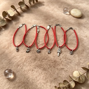 Könnte beinhalten: Sechs rote Kordelarmbänder, jedes mit einem einzigartigen silberfarbenen Metallanhänger. Die Anhänger zeigen einen kleinen Schuh, eine Lippenform, ein Herz, einen Stern, ein Schwert und eine kleine Figur. Jedes Armband hat ein kleines silbernes Etikett.