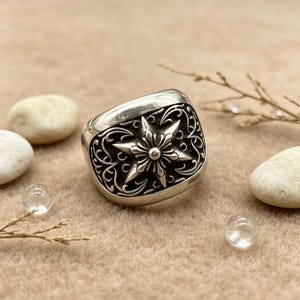 Könnte beinhalten: Ein silberner Ring mit einem markanten sechsstrahligen Stern in der Mitte, umgeben von komplizierten, dunkel oxidierten Rankenmustern. Die Metalloberfläche ist poliert und hebt das detaillierte Relief des Sterns sowie die wirbelnden Filigranmotive hervor, die sich über die Vorderseite des Rings erstrecken.