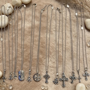 May include: A collection of ten silver-toned metal necklaces arranged in a row. Each necklace features a distinct pendant, including various cross designs, a circular emblem, and a rectangular tag, all suspended from thin, delicate link chains.