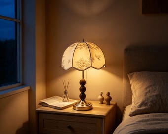 May include: A table lamp with a scalloped, off-white fabric shade featuring a delicate grey leaf pattern. The lamp base consists of three stacked dark brown wooden spheres atop a circular metallic stand, emitting a warm, soft glow in a room.