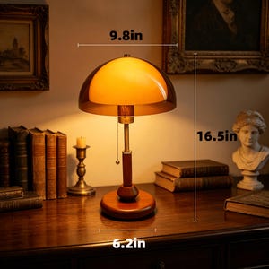 Puede incluir: Una l&aacute;mpara de mesa con pantalla de cristal &aacute;mbar en forma de c&uacute;pula, v&aacute;stago color lat&oacute;n y base redonda de madera. La l&aacute;mpara cuenta con un interruptor de cadena y mide 24,9 cm de ancho, 41,9 cm de alto y 15,7 cm de di&aacute;metro de base.