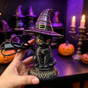 May include: A black cat figurine wearing a purple witch hat with a gold buckle. The cat has yellow eyes, a crescent moon charm on its chest, and sits on a decorative base. A black power cord extends from the side of the cat figure.