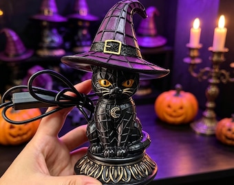 May include: A black cat figurine wearing a purple witch hat with a gold buckle. The cat has yellow eyes, a crescent moon charm on its chest, and sits on a decorative base. A black power cord extends from the side of the cat figure.