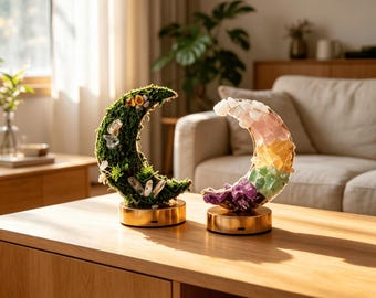 May include: Two crescent moon-shaped decorative lamps on gold circular bases. The left lamp is covered in green moss with embedded clear crystals. The right lamp features a gradient of multi-coloured raw crystals including purple, green, yellow, and pink.