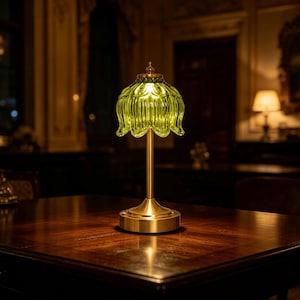 Puede incluir: Una lámpara de mesa con una pantalla de vidrio estriado de color verde lima translúcido en forma de pétalo de flor. La lámpara tiene un tallo de metal fino de color latón cepillado y una base circular, emitiendo un brillo suave y cálido.